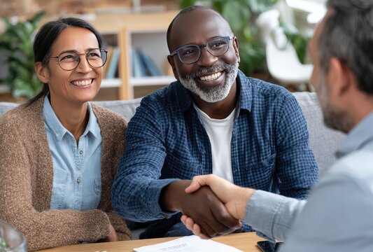 A smiling interracial couple shakes hands with another person, likely sealing a deal or agreement, in a bright, neutral, home-like setting