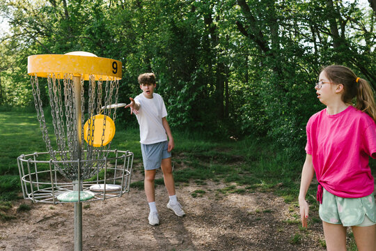 Siblings throw discs into golf basket outside in green forest