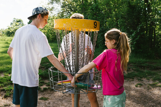 Siblings pick up discs from golfing basket in green forest sunshine