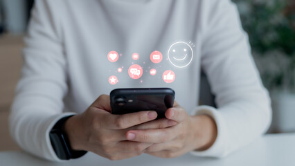 Businesswoman holding smartphone with hologram social media icons such as like, chat, email, and smiley face. Concept of online communication, customer engagement, and digital marketing.