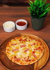 Tasty pineapple and ham hawaiian pizza flatbread baked on wooden plate, delicious melting mozzarella cheese on wooden board, homemade cooking ingredients lying on dark background, food advertisement.