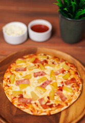 Tasty pineapple and ham hawaiian pizza flatbread baked on wooden plate, delicious melting mozzarella cheese on wooden board, homemade cooking ingredients lying on dark background, food advertisement.