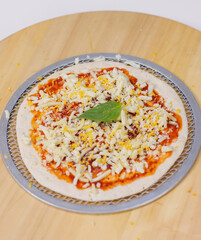 Unbaked tasty cheese crispy pizza flatbread baked on baking steel plate, delicious melting mozzarella cheese on wooden board, homemade cooking ingredients lying on light background, food advertisement