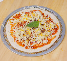 Unbaked tasty cheese crispy pizza flatbread baked on baking steel plate, delicious melting mozzarella cheese on wooden board, homemade cooking ingredients lying on light background, food advertisement