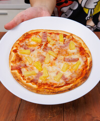 Tasty pineapple and ham hawaiian pizza baked on baking steel plate, delicious melting mozzarella cheese on wooden board, homemade cooking ingredients lying on dark background, food advertisement.