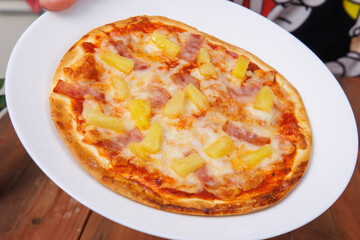 Tasty pineapple and ham hawaiian pizza baked on baking steel plate, delicious melting mozzarella cheese on wooden board, homemade cooking ingredients lying on dark background, food advertisement.