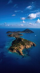 Stunning aerial view of a rugged, narrow island in a vast, deep blue sea under a bright blue sky with scattered clouds on a sunny day, showcasing nature's beauty
