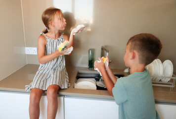 Smiling children playing with sponge and soap foam while washing dishes at kitchen sink