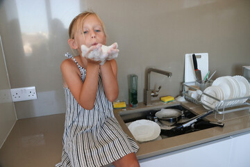 Smiling little girl playing with soap foam while helping with dishwashing at kitchen sink