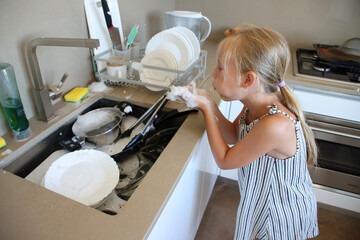 Little girl blowing soap foam bubbles while helping with dishwashing in bright kitchen