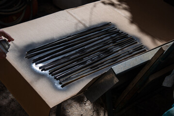 Hand spray painting a set of black metal rods on cardboard surface in sunlight, close-up view