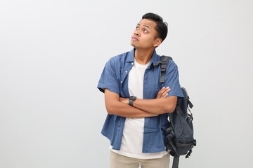 Fototapeta premium Young asian student man wearing casual clothes and backpack, standing with folded arms and looking up in empty space, isolated on white background.