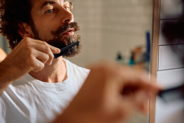 Reflection in mirror of man grooming his beard in bathroom.