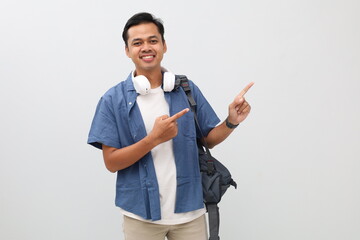 Young Asian male student wearing casual clothes and backpack, headphones around neck, pointing up at empty space, presenting something, standing smiling looking at camera, isolated white background.