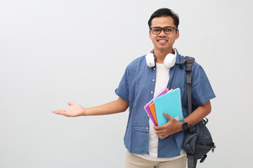 Young Asian male student wearing casual clothes, backpack, glasses, headphones around neck and carrying books, making a gesture of extending his hand to the side in empty space, inviting.