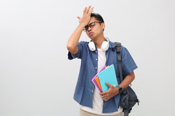 Young Asian male college student wearing casual clothes, backpack, glasses, headphones around neck while carrying books, forgetting something or making a mistake while holding forehead.