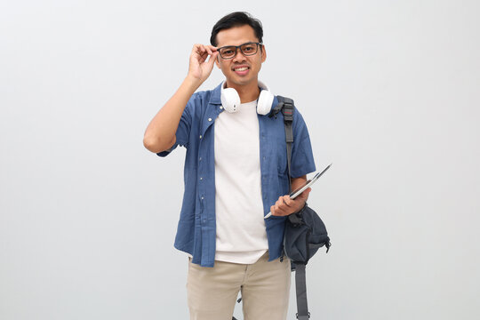 Young asian male student wearing casual clothes, backpack, headphones around neck, carrying digital tablet computer, holding glasses, looking at camera while smiling, isolated white background. - Powered by Adobe