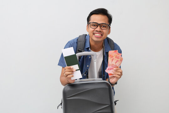 An asian traveler man wearing casual clothes, holding a fan filled with cash rupiah banknotes, suitcase and passport ticket, air flight travel, tourism or vacation concept.