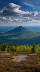 A conical green mountain rises above a treeline under a bright, partially cloudy, blue sky, with a rocky plateau foreground creating a soft-focus effect