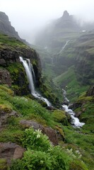 Obraz premium A scenic waterfall cascading down a mossy cliff into a stream winding through a lush green valley, with a misty mountainous backdrop under an overcast sky