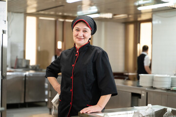 Portrait of a female chef in a black uniform with red details smiling in a professional restaurant kitchen. Image highlights hospitality staff, confidence, and culinary professionalism.