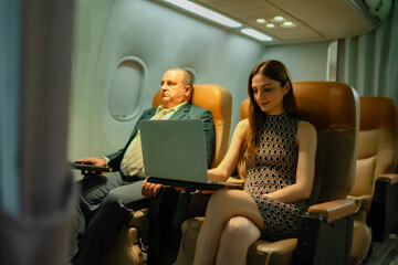 Fototapeta premium Smiling overweight businessman seated next to woman working on computer during flight. Idea of inflight productivity, business trip, technology use, airline service, and international travel industry.