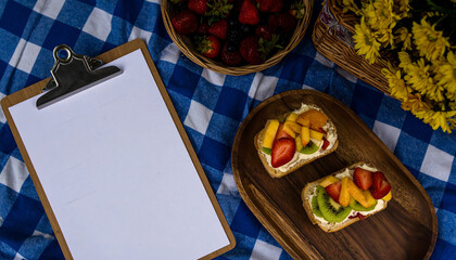 Blank restaurant menu clipboard styled with fruit cream sandwiches and flowers picnic setup. AI
