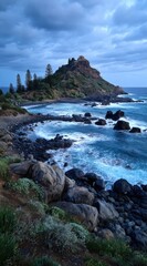 Coastal landscape features a rocky shoreline, dynamic ocean waves, a small island, and a cloudy sky at dusk