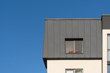 Detail of the upper floor of a modern building against a clear blue sky. There is a grey roof, and red flowers are visible on the window. Background.