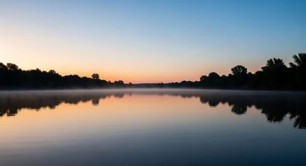 Obraz premium A tranquil sunrise over a misty lake, with the colorful sky and silhouetted trees perfectly reflected in the calm, glassy water.