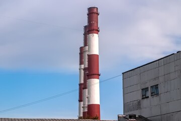 Tall red and white industrial chimneys rise against a partly cloudy sky, adjacent to a grey,...