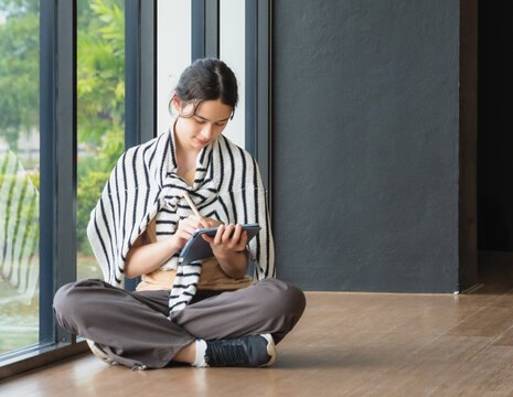 Young woman sits on floor next large window, writing tablet. wear sweater. greenery outside window creates calm and focus study environment, ideal for portraying self-learning knowledge  education.