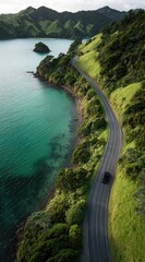 A winding coastal road cuts through lush green hills, bordering a tranquil turquoise bay, a small dark vehicle making its way along the curved asphalt