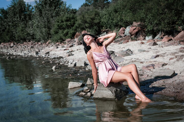 Fototapeta premium Young sexy beautiful woman is sitting on the rocks in the sea near the shore and enjoys relaxation, the warm sun and nature. Wellness, tourism, holidays, vacation and weekends. 