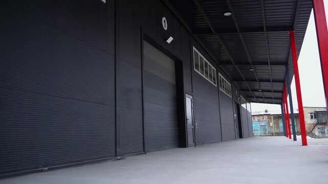 Empty ramp of large industrial complex with black facade and poles. Minimalist warehouse exterior with closed gates and doors on gloomy day