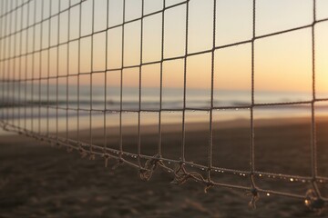 Beach volleyball net glistens with morning dew as sun sets over calm ocean waves