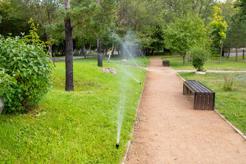 Sprinklers water the lush green grass in a peaceful park on a sunny day. Walkways lined with benches. renovation of urban environment