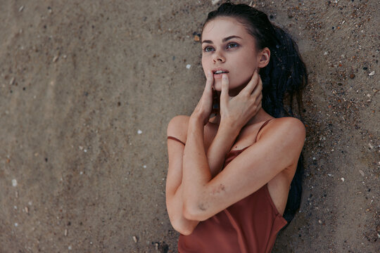 Young woman lies on a rough wall, hands near her mouth, with a pensive gaze and subtle vulnerability. Warm tones, soft light, and an intimate, candid mood define the frame.