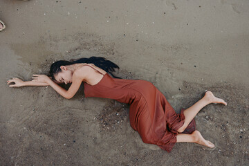 Woman lying on sandy ground in a flowing red dress, relaxed pose with arms extended, dramatic composition capturing texture, warmth, and subtle motion on a sunlit beach.