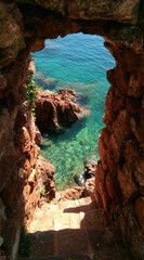 A stone archway frames a view of turquoise sea, rocks, and steps descending downwards. Sunlight illuminates the scene, creating a sense of peace