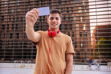 Portrait of a smiling young man with red headphones around his neck showing a card in the...