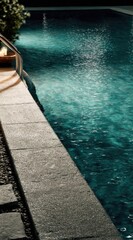 A tranquil nighttime shot of a turquoise swimming pool with stone coping. The water reflects the surrounding lights, creating a shimmering effect