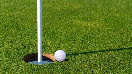 A white golf ball sits on the lip of the cup on a perfect green putting surface with a flagstick