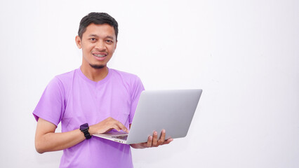 Happy asian man wearing purple t-shirt and holding laptop computer working online and looking at camera isolated on white background, online education