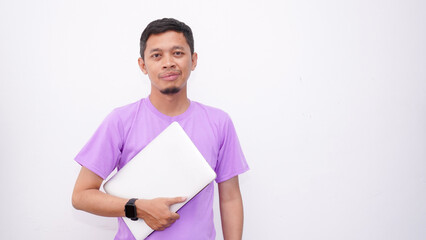 Happy asian man wearing purple t-shirt and holding laptop computer working online and looking at camera isolated on white background, online education