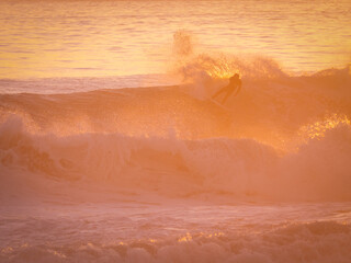 Fototapeta premium SILHOUETTE: Unknown surfer carves along the face of a breaking wave, backlit by intense golden light of setting sun. As he makes a top turn, water sprays around. Riding waves at famous Moroccan spot.