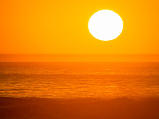 LENS FLARE, CLOSE UP: Sun hangs over the ocean horizon, casting an intense warm orange glow. Water and sky are a monochromatic study in gold, emphasizing dramatic beauty of evening light in Morocco.