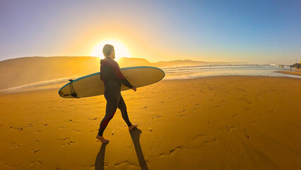 LENS FLARE, PORTRAIT: Surfer walks along wide, golden beach at sunrise, with his surfboard tucked...