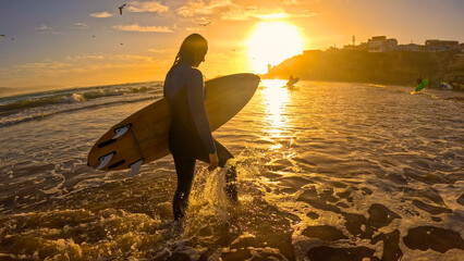 LENS FLARE: Female surfer with wooden surfboard wades through shallows at sunset. She came out of water after evening surf session and is walking along the shoreline, bathed in golden setting sun.