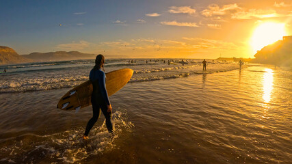 LENS FLARE, SILHOUETTE: Female surfer in wetsuit walks along scenic ocean shore at sunset after evening session. She is carrying surfboard as she wades in shallow water at famous Moroccan surf break.
